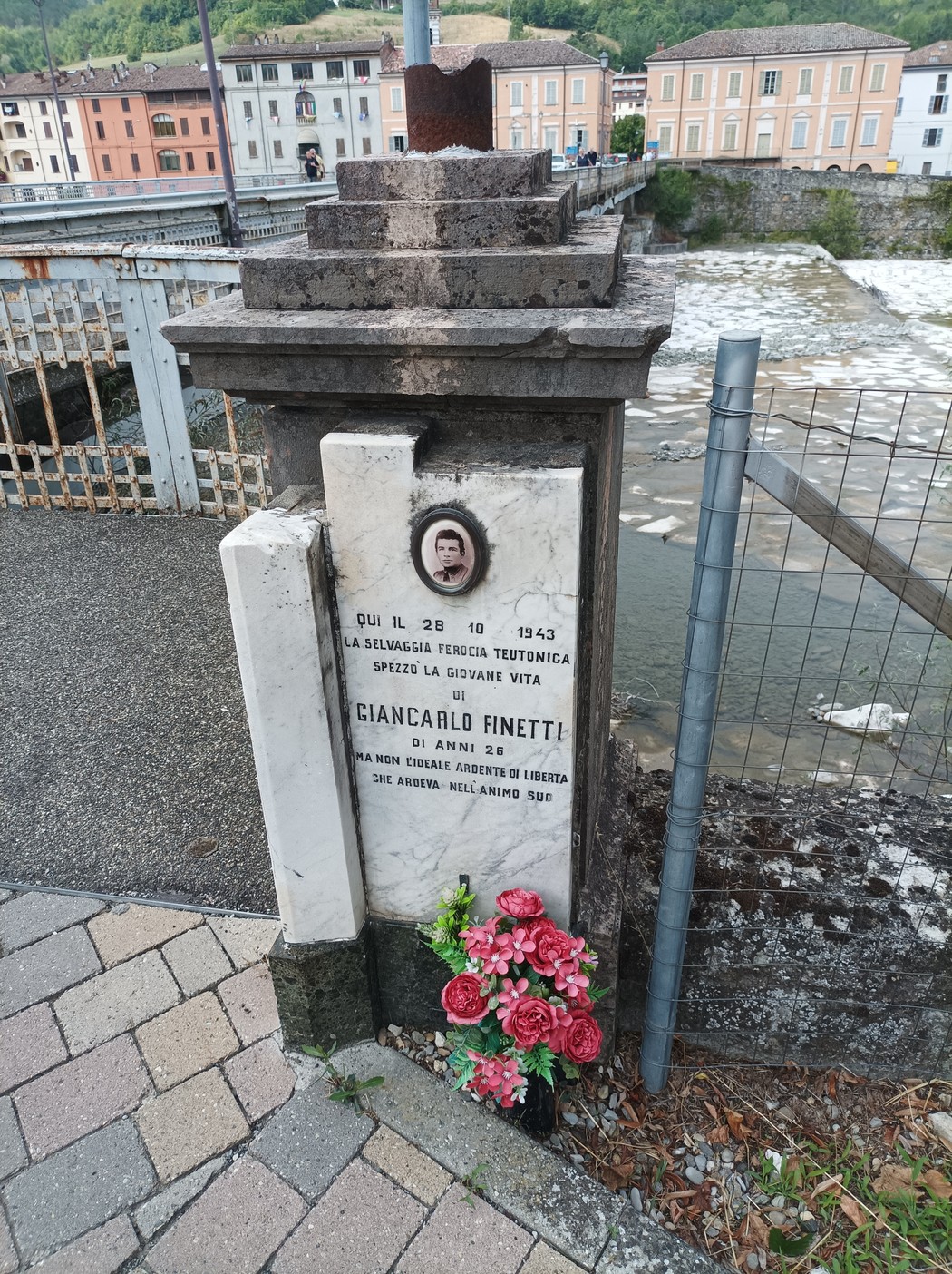 Cippo commemorativo di Cianalbi Finetti, con fiori e foto, sullo sfondo il fiume e case storiche.