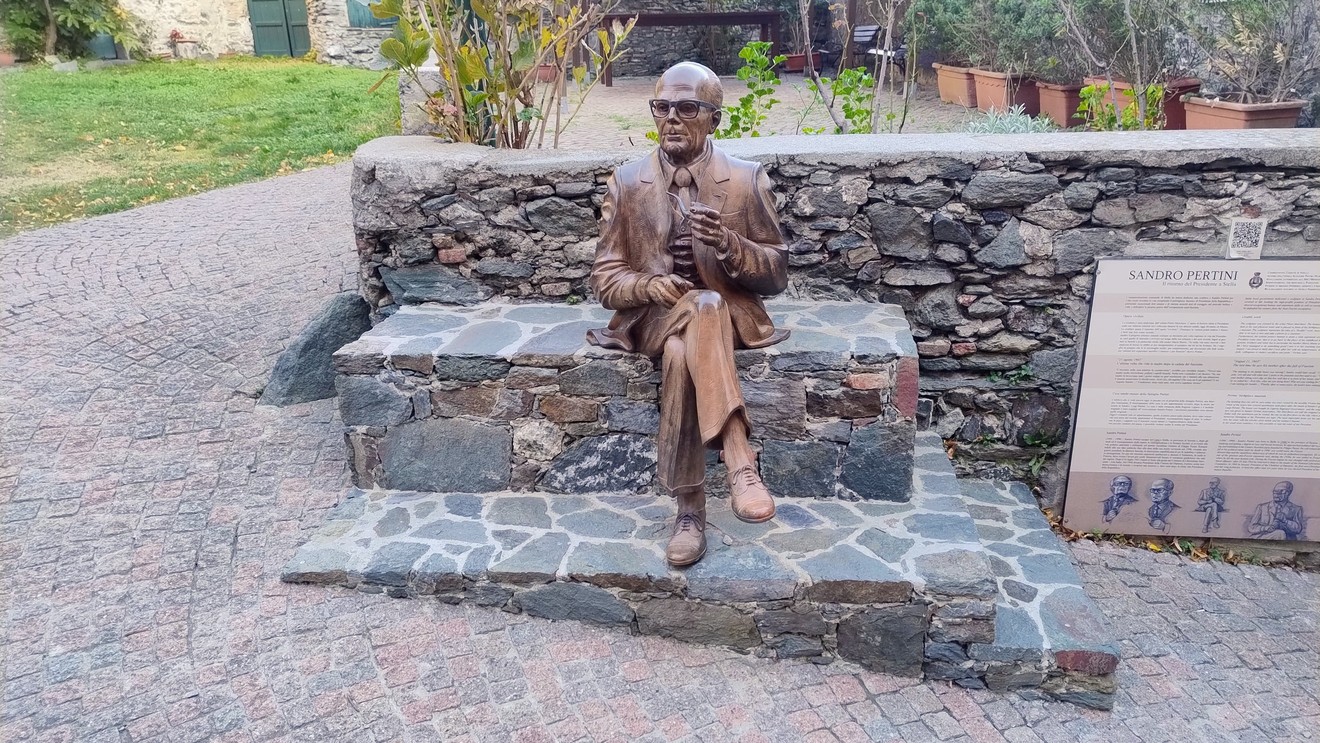 Statua bronzea di Sandro Pertini seduto su una panchina in un contesto urbano.