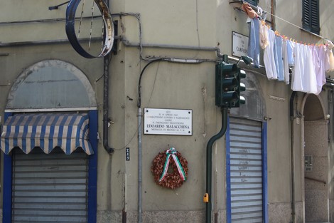 Lapide commemorativa dedicata a Edoardo Malacchina su una facciata di un edificio, con strisce di biancheria stesa.