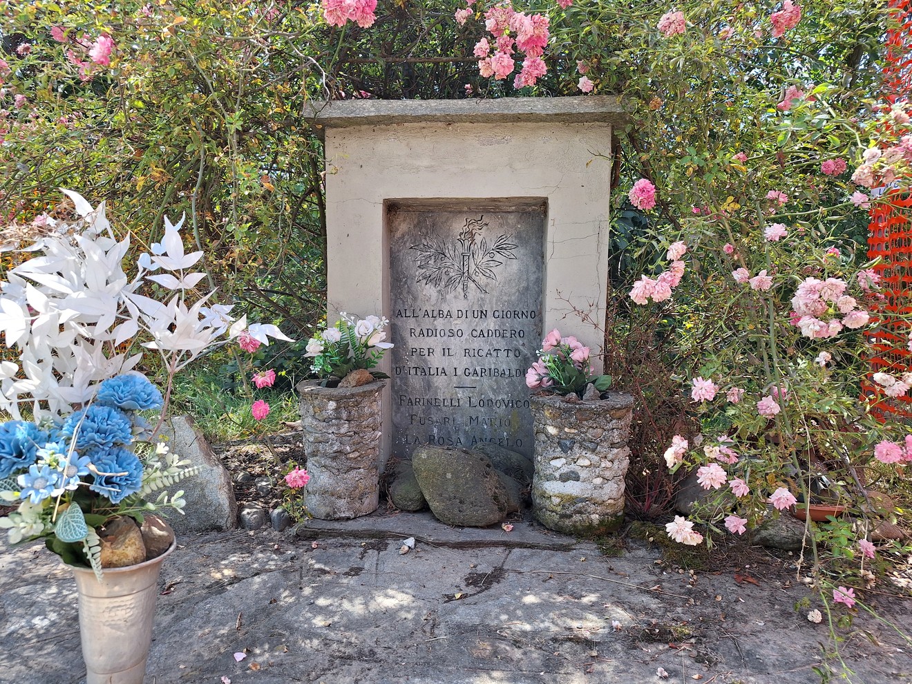 Cippo commemorativo circondato da fiori, dedicato a Farinelli, Fusari e La Rosa, con decorazioni floreali.
