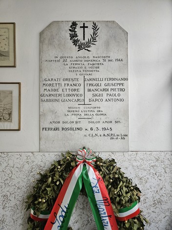 Lapide in marmo con croce scolpita e iscrizione, appesa a parete, sotto una corona floreale con nastro tricolore.