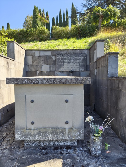 Sepolcro in pietra con lapide incisa, immerso in un giardino cimiteriale con siepi e cipressi sullo sfondo.