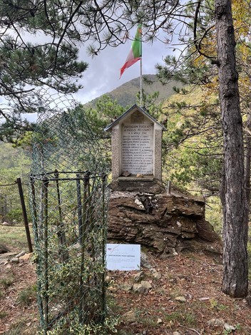Monumento commemorativo ai Martiri di Passo Mezzano, circondato da alberi e natura, con bandiera italiana.