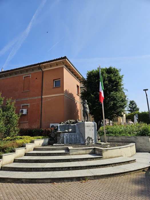 Monumento ai Caduti a San Rocco al Porto, con scalinata, bandiera italiana e sfondo di edifici e verde.