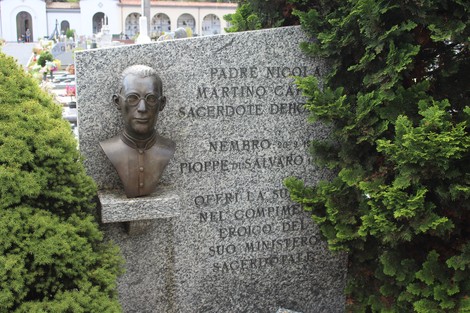 Monumento memoriale con busto e dedica per i caduti, circondato da piante verdi in un contesto cimiteriale.