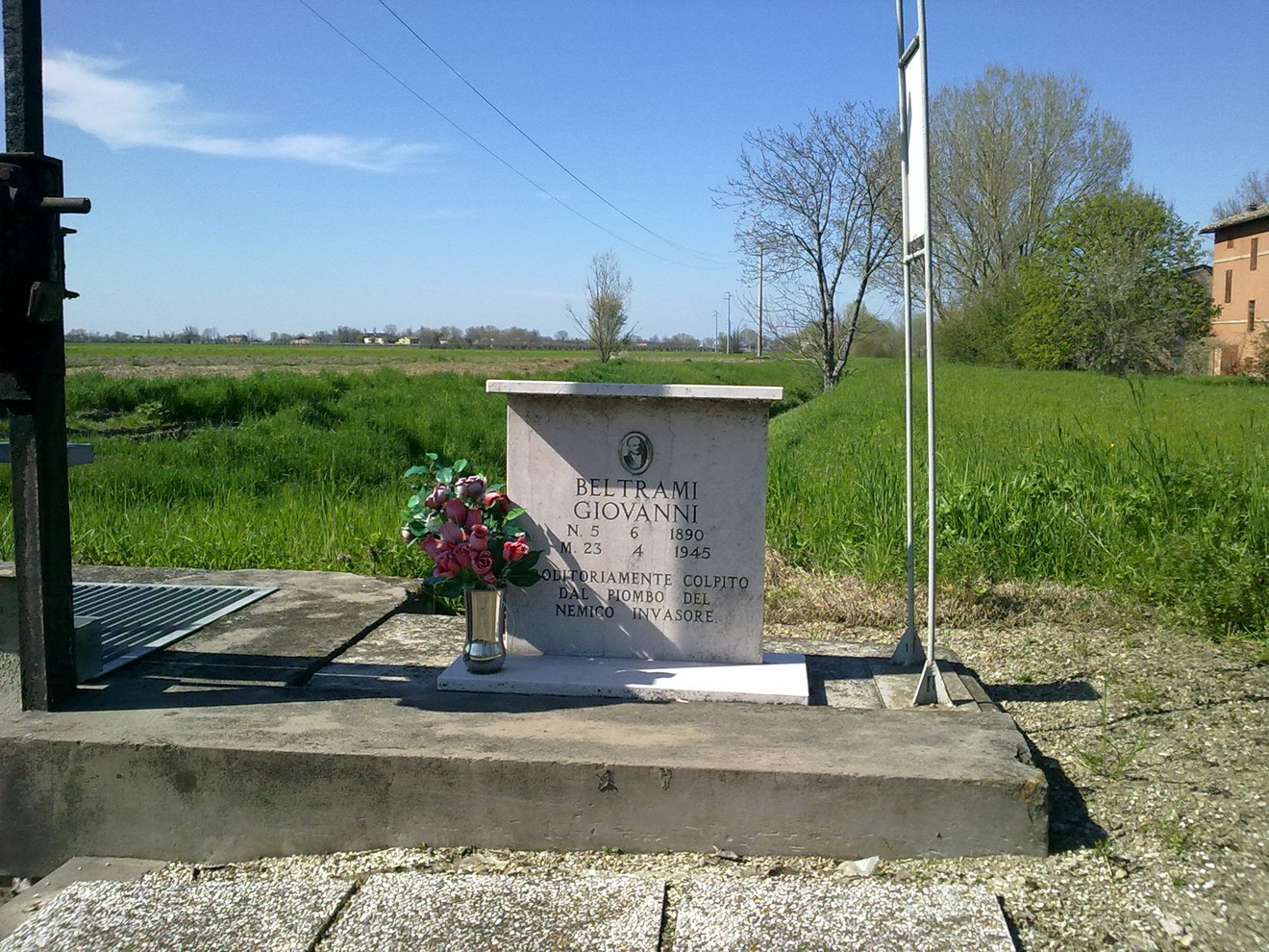 Cippo commemorativo a Beltrami, con fiori e il paesaggio rurale sullo sfondo.