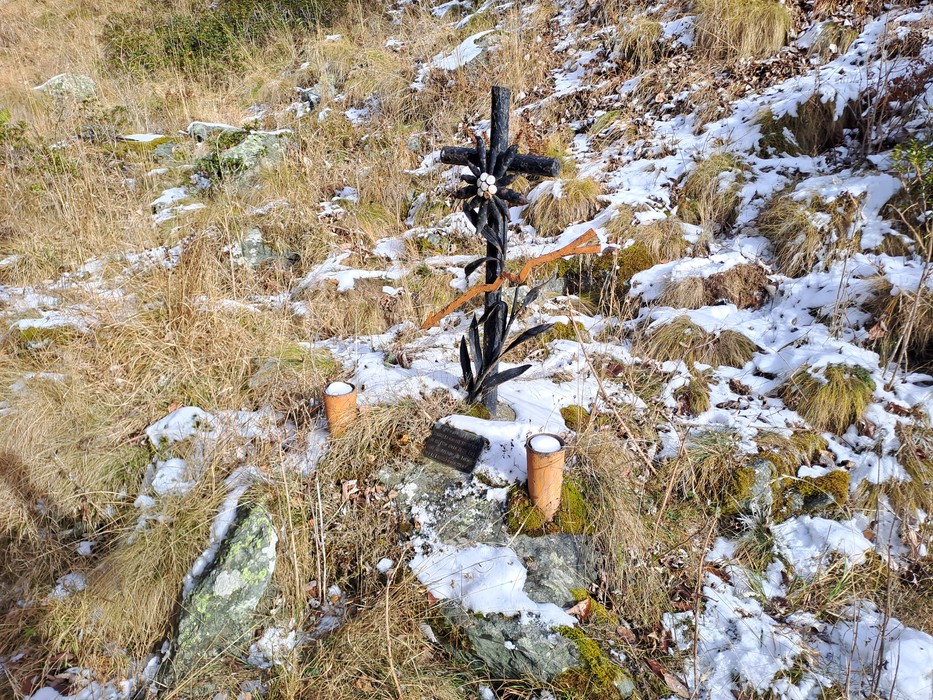 Croce in ferro battuto su roccia tra erba e neve, con targa incisa e due candele ai lati.