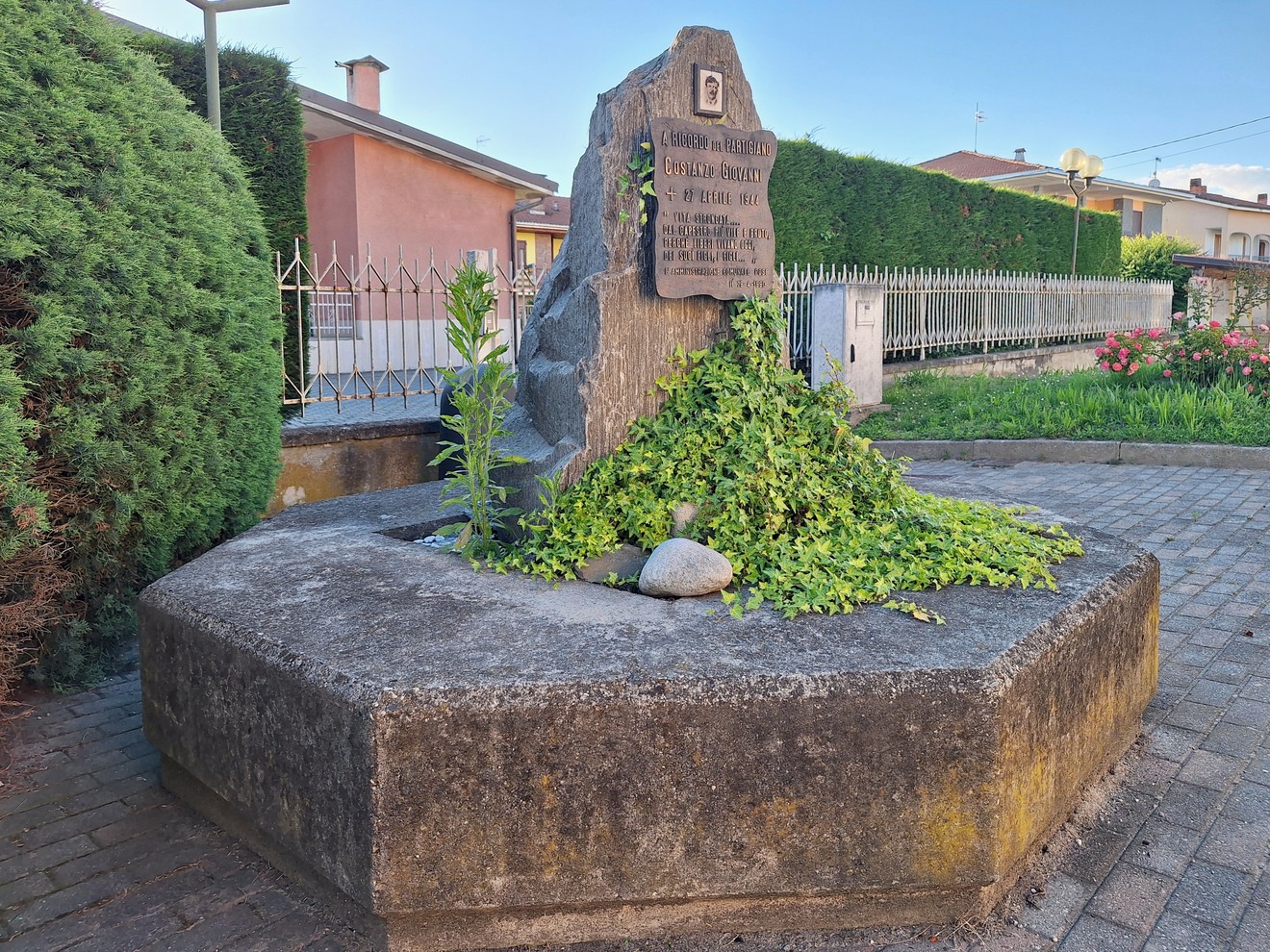 Cippo commemorativo ricoperto di vegetazione, situato in un'area verde con case sullo sfondo.