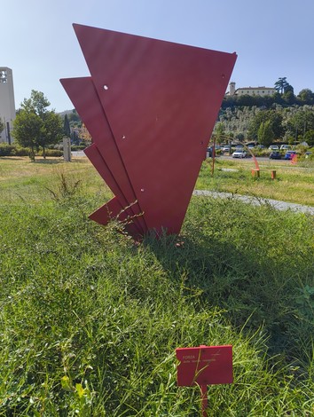 Scultura rossa a forma di triangoli sovrapposti immersa in un prato verde, in un contesto urbano.