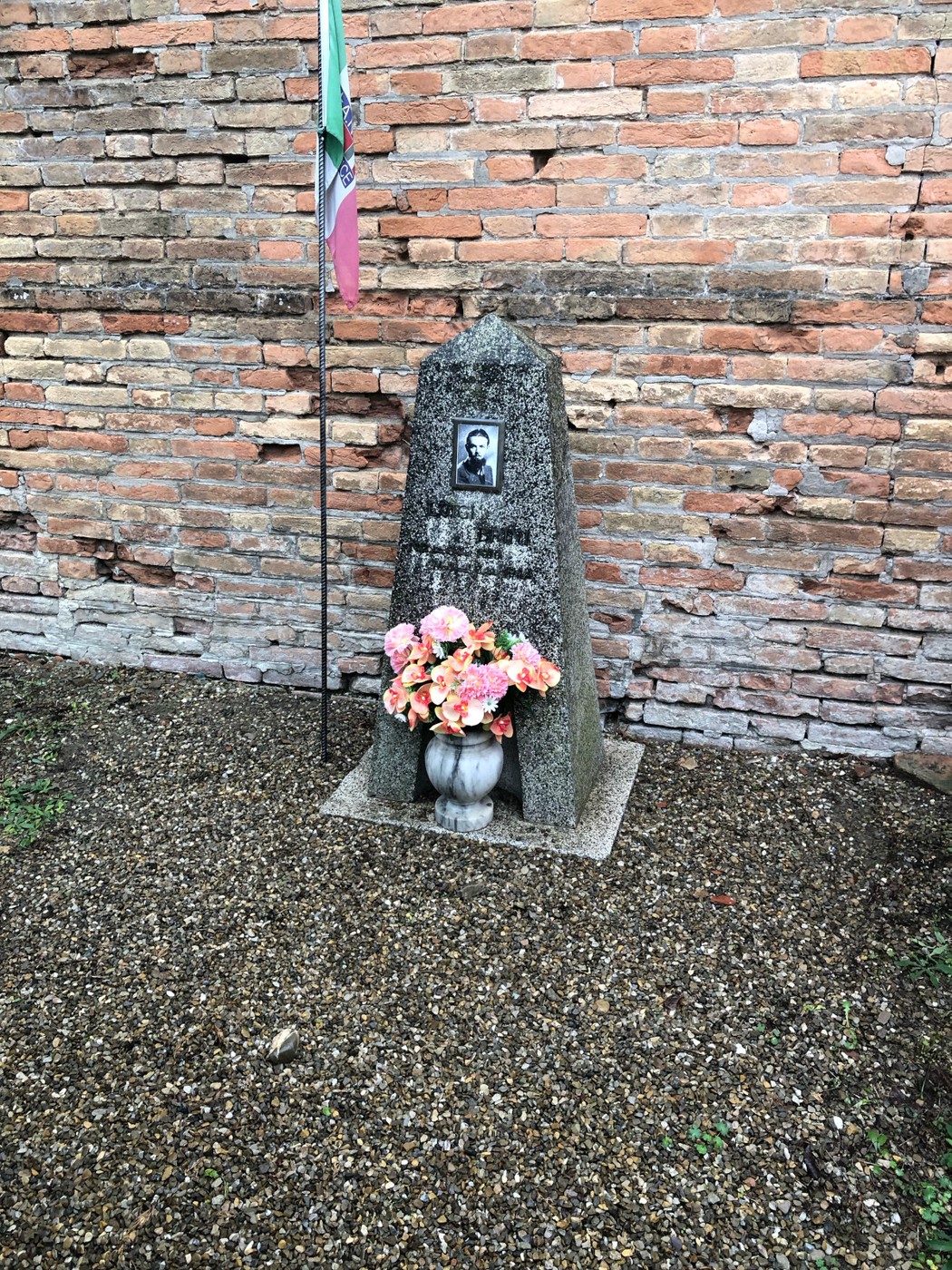 Cippo a Brini con fiori e bandiera, situato davanti a un muro di mattoni.