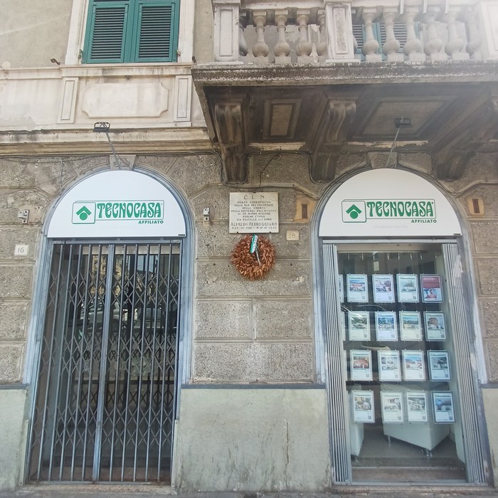 Lapide con iscrizione tra due archi di un'agenzia Tecnocasa, sulla facciata di un edificio storico.