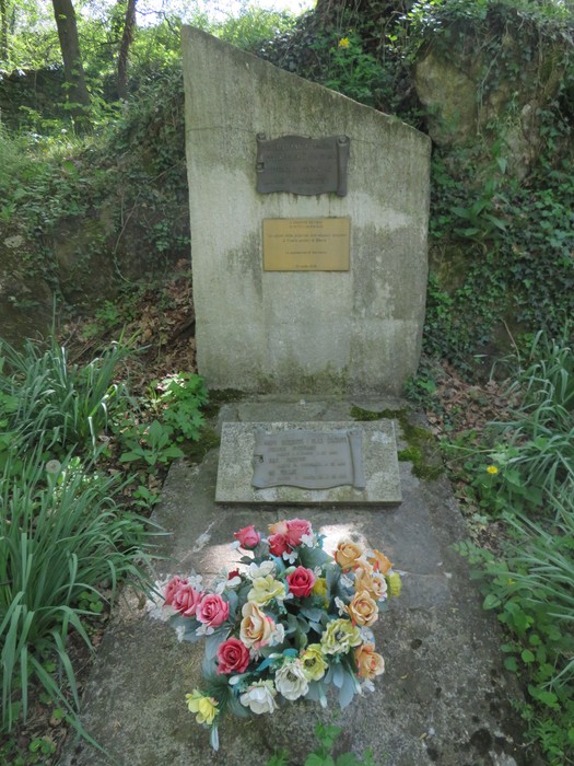 Monumento commemorativo nel Parco della Rimembranza, decorato con fiori artificiali e circondato dalla vegetazione.