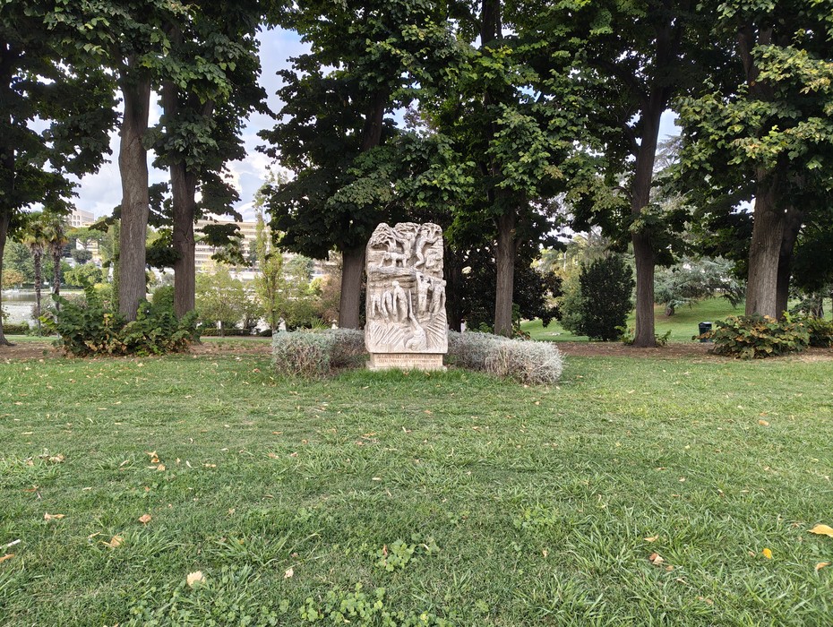 Monumento in pietra con rilievi e iscrizione, collocato su un prato alberato.