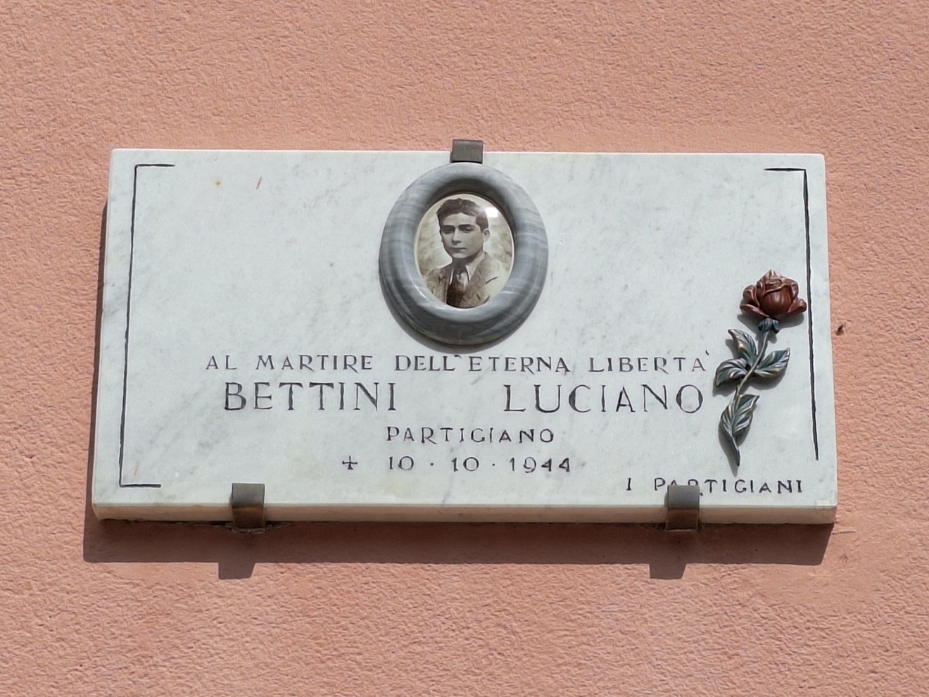 Lapide commemorativa dedicata a Luciano Bettini, partigiano, con foto e decorazioni floreali.