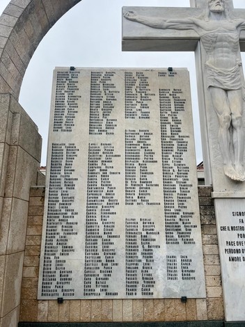 Monumento commemorativo con una grande lastra di pietra, nomi incisi, e una croce stilizzata sullo sfondo.