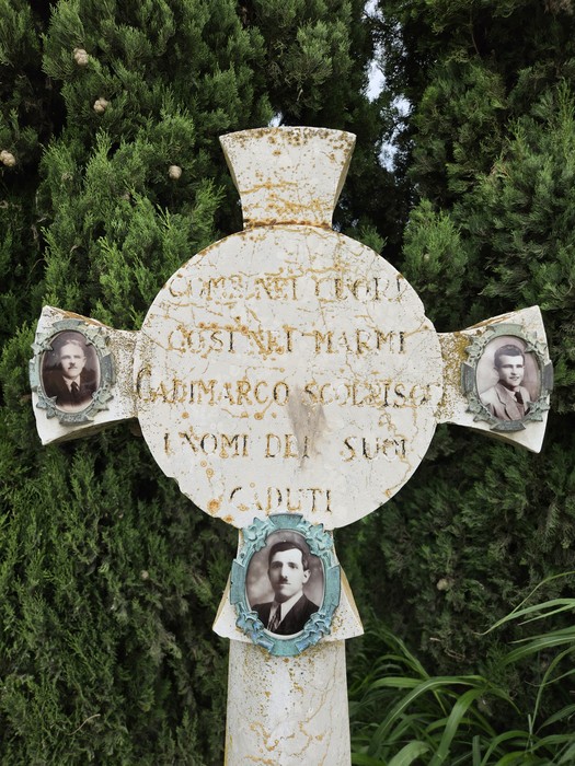 Cippo commemorativo con croce e fotografie, immerso nel verde, dedicato ai caduti.