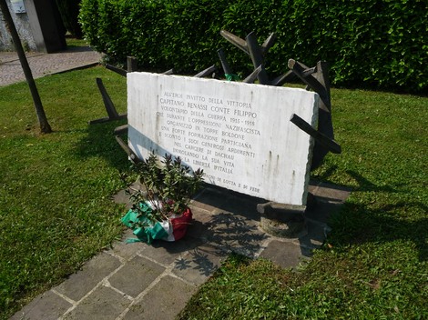 Lapide commemorativa dedicata a Benassi, con fiori e drappo tricolore sullo sfondo di un giardino.