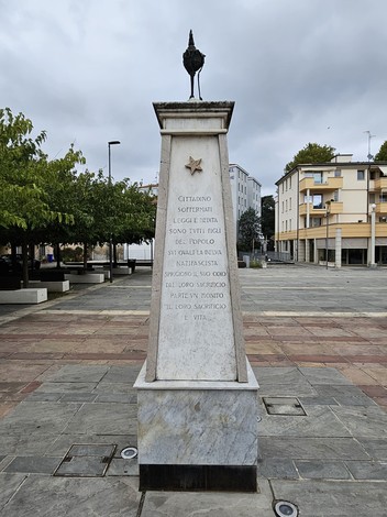 Monumento ai Partigiani con iscrizioni commemorative, situato in uno spazio pubblico con alberi e edifici circostanti.