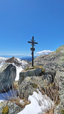 Croce di metallo posta su un masso, circondata da paesaggio montano con neve e cielo azzurro.
