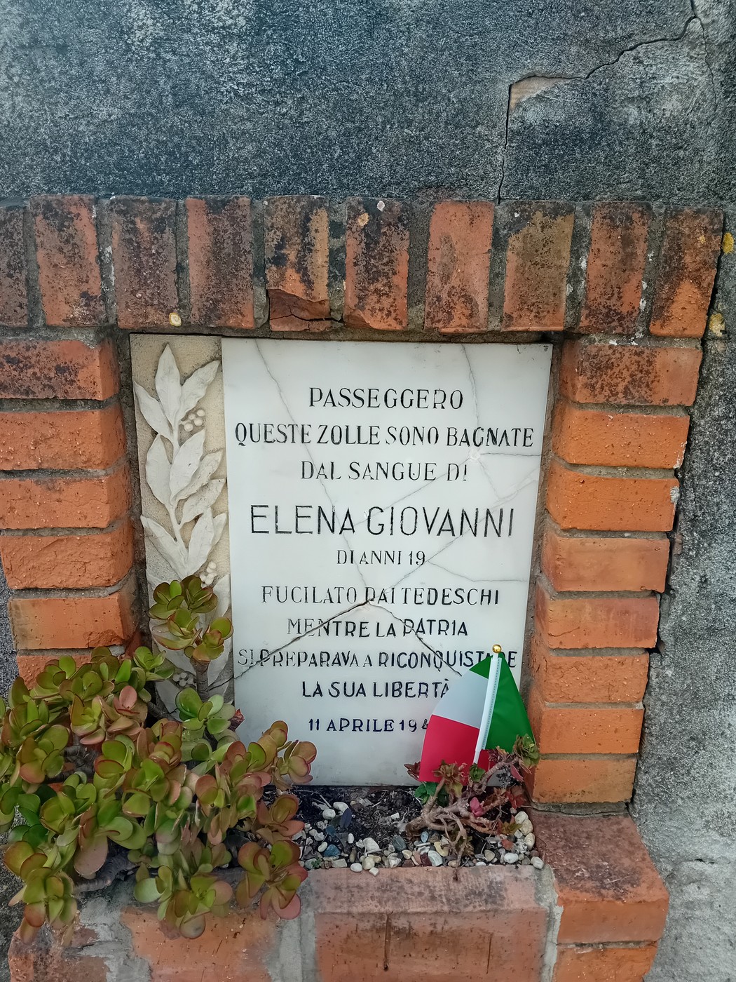 Lapide commemorativa dedicata a Elena Giovanni, con piante e bandiera italiana.