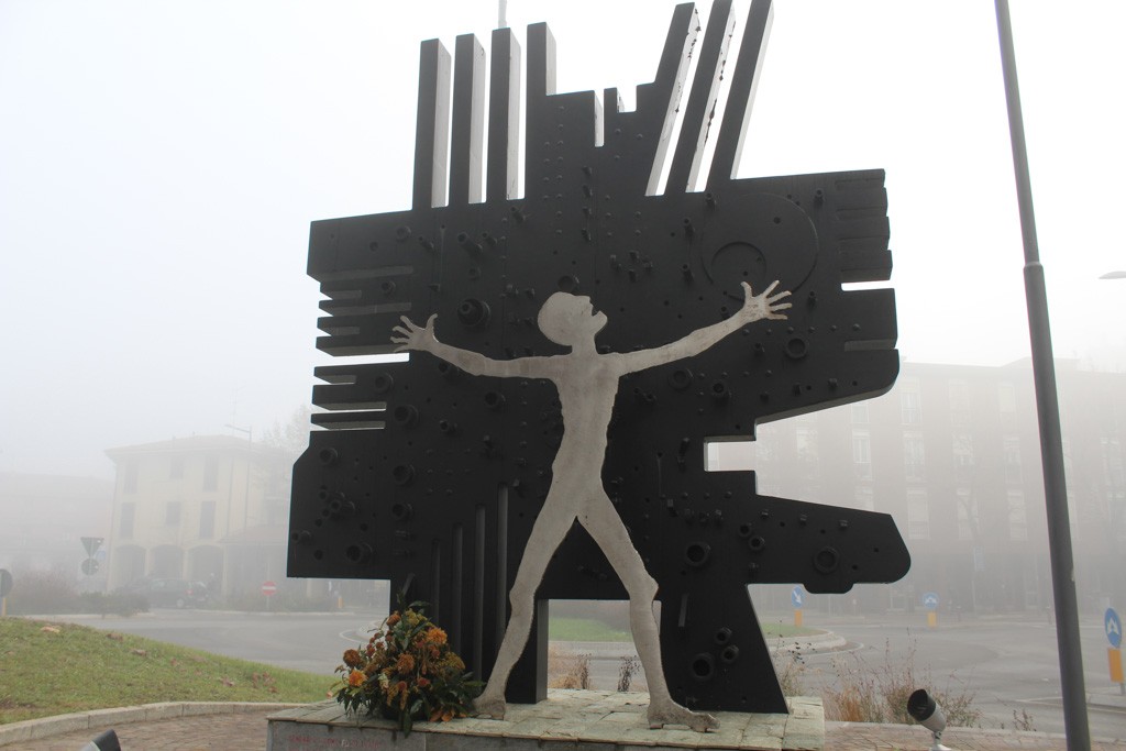 Monumento in metallo scuro con sagoma umana stilizzata a braccia aperte, in una piazza con fiori deposti alla base.