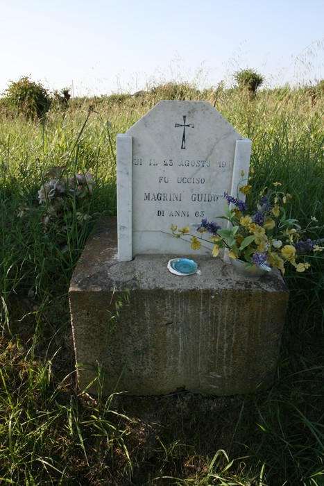 Cippo commemorativo dedicato a Guido Magrini con fiori e croce, situato in un ambiente naturale.