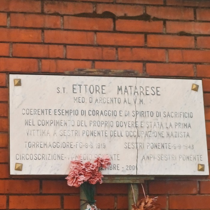 Lapide marmorea con iscrizione su muro di mattoni, decorata con garofani rosa in un vaso