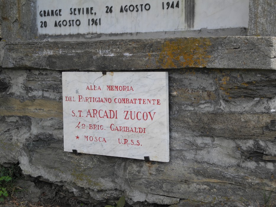 Targa in marmo con iscrizione, fissata a un muro in pietra, sotto una lapide più grande.