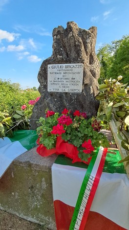 Cippo commemorativo dedicato a Giulio Biscazzo, adornato da fiori e bandiere italiane.