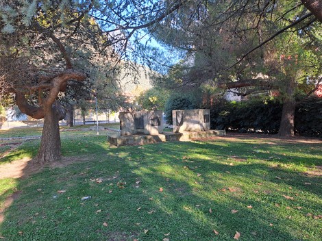 Monumento ai Caduti in un parco, circondato da alberi e prato verde, con due lastre commemorative visibili.