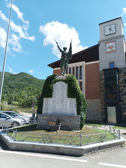 Monumento ai caduti con statua alato, circondato da vegetazione e edifici, in un paesaggio montano.