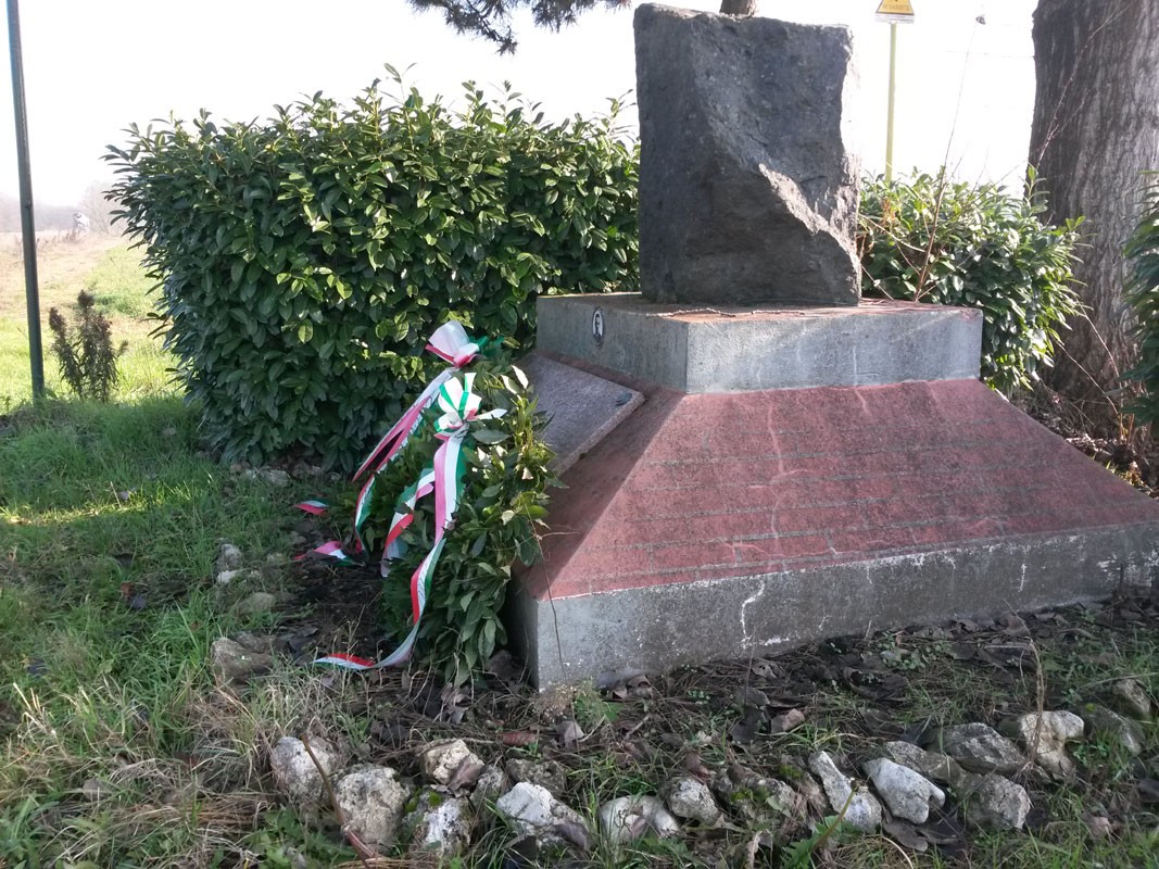Monumento a Muzio, con base in pietra e decorazioni tricolori, immerso nel verde.