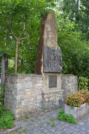 Monumento a Ricci, incastonato nel verde, caratterizzato da una struttura in pietra e rilievi artistici.