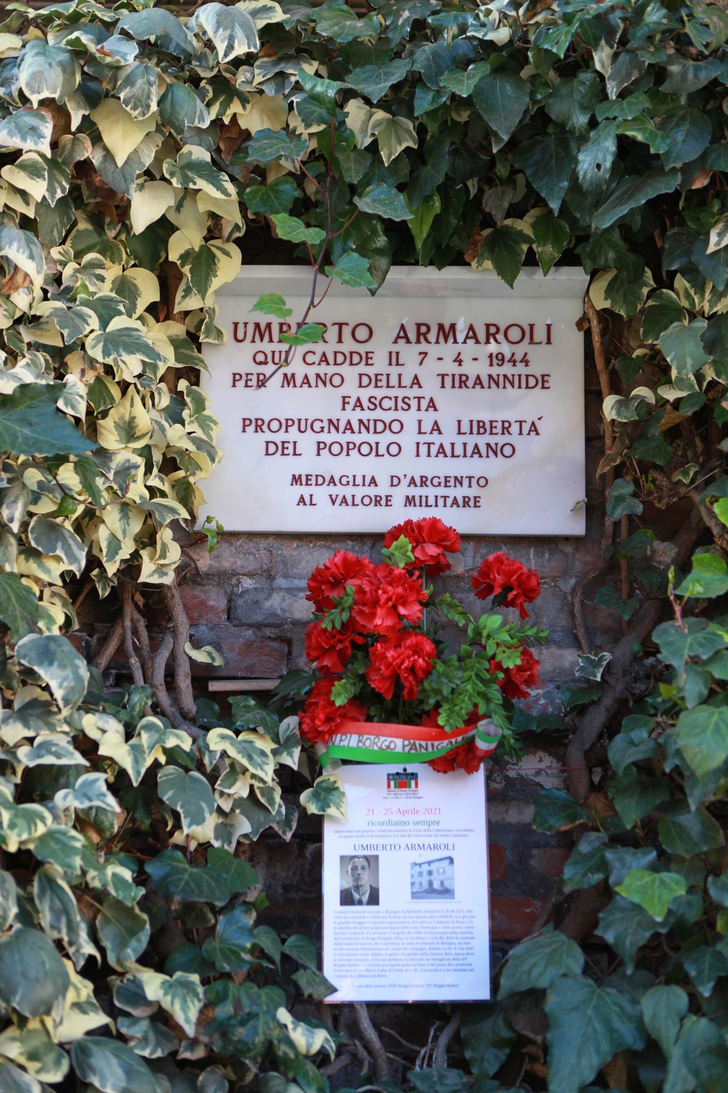 Lapide commemorativa di Umberto Armaroli, decorata con fiori rossi e circondata da edera.