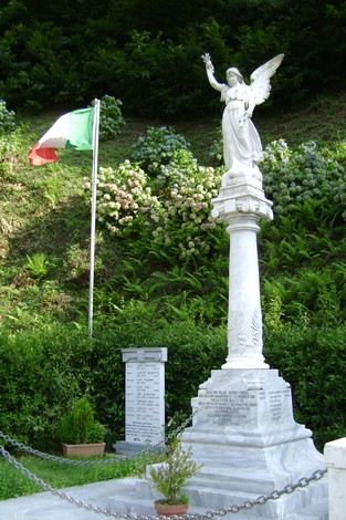 Monumento ai caduti con statua di un angelo, circondato da verde e bandiera italiana. Riflessione e memoria.