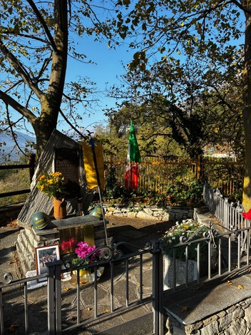 Monumento con targa incisa, bandiera tricolore e fiori, recintato da staccionata in metallo tra gli alberi.