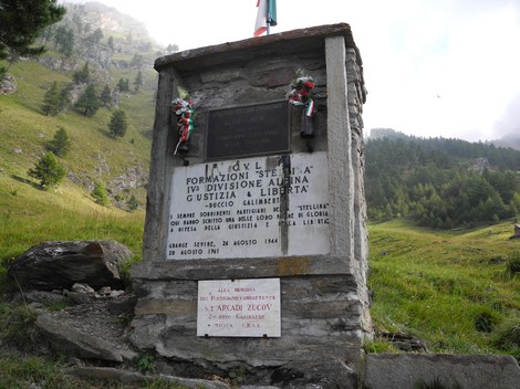 Cippo in pietra con lapidi e fiori, sormontato da bandiera, su prato tra alberi e montagne.