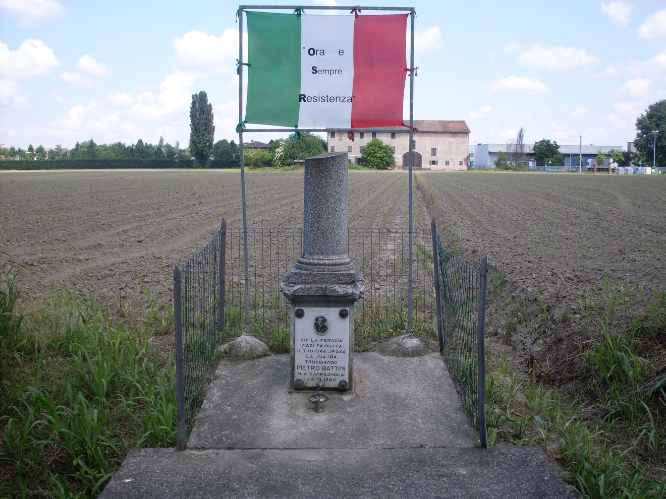 Cippo commemorativo a Battini, con bandiera italiana e messaggio di resistenza. Circondato da un campo aperto.