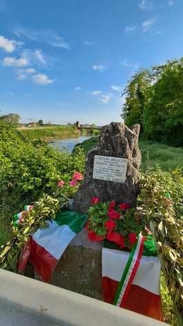 Cippo commemorativo decorato con fiori e bandiere, immerso nella natura vicino a un fiume.
