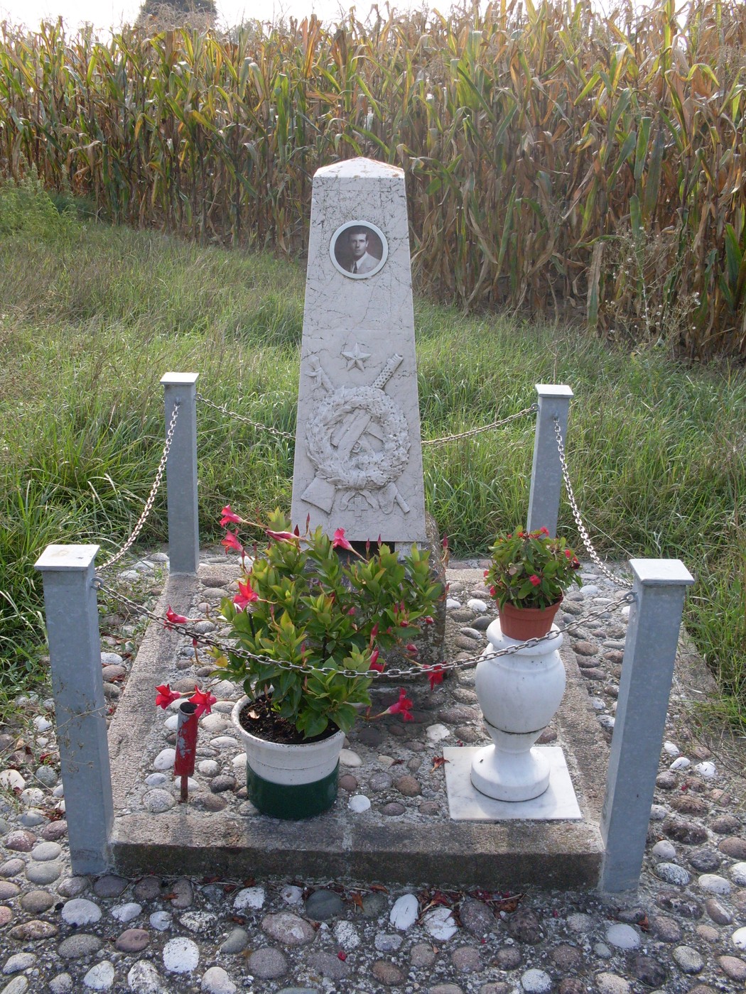 Cippo commemorativo con fiori, situato in un campo, dedicato alla memoria di una figura storica.