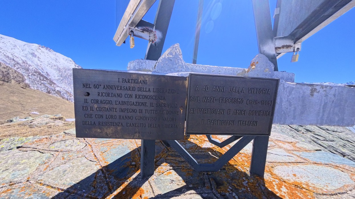 Due targhe metalliche con iscrizioni su supporto in quota, montagne innevate sullo sfondo.