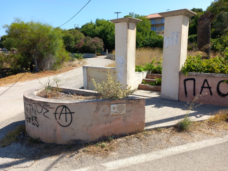 Lapide commemorativa in un'area rurale, circondata da vegetazione e segni di degrado urbano.