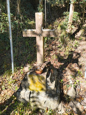 Croce commemorativa nel verde, circondata da foliage e segni di usura, simbolo di memoria e venerazione.