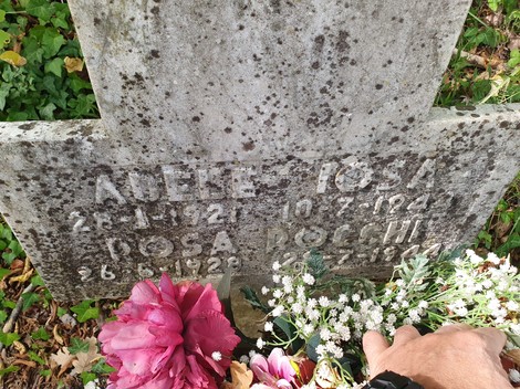Croce funeraria con nomi e date, decorata con fiori, in un ambiente naturale.