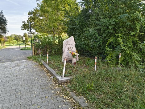 Cippo commemorativo immerso nel verde, circondato da fiori e segnaletica stradale.