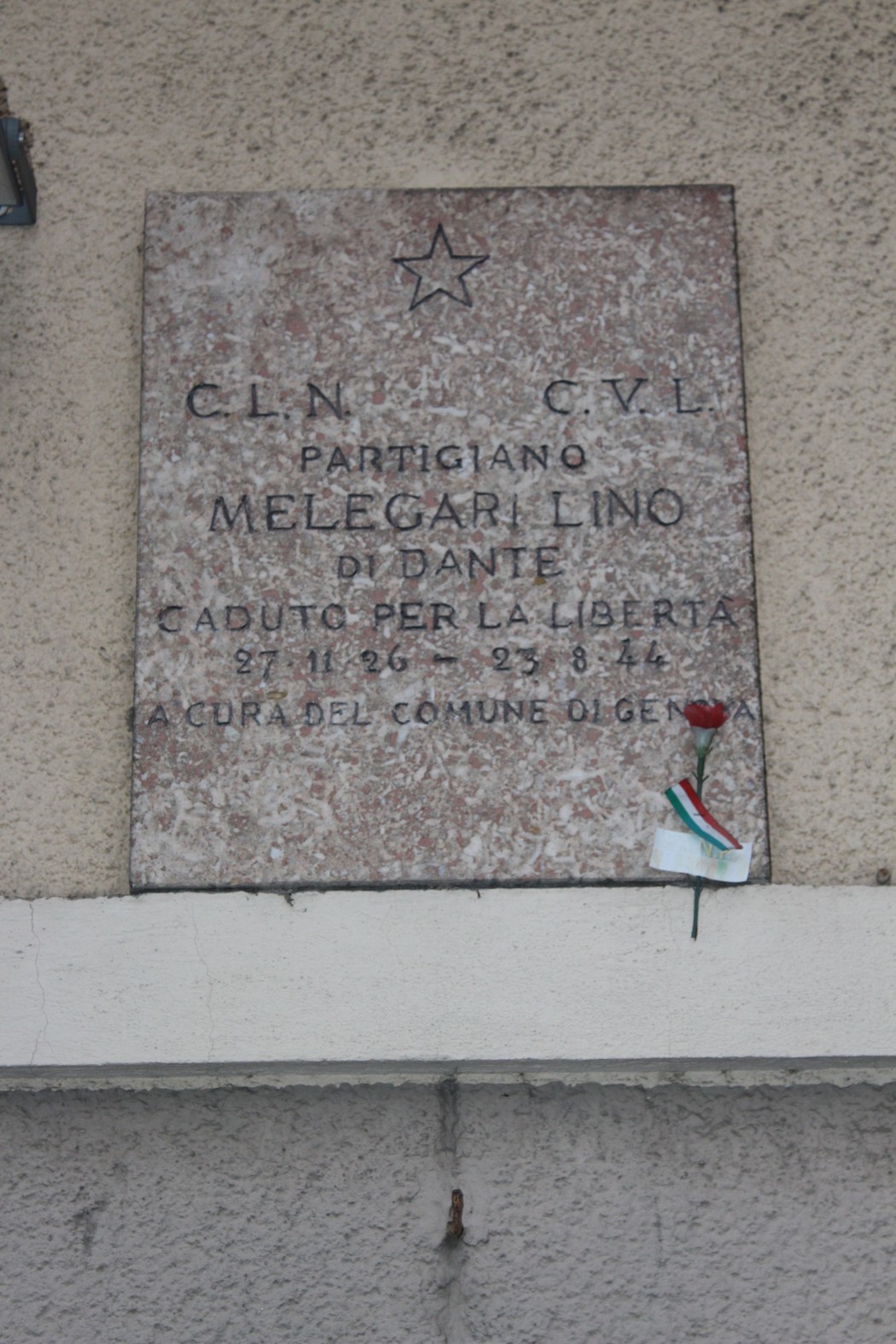 Lapide commemorativa in onore del partigiano Melegari Lino, caduto per la libertà. Decorata con un fiore e una bandiera.