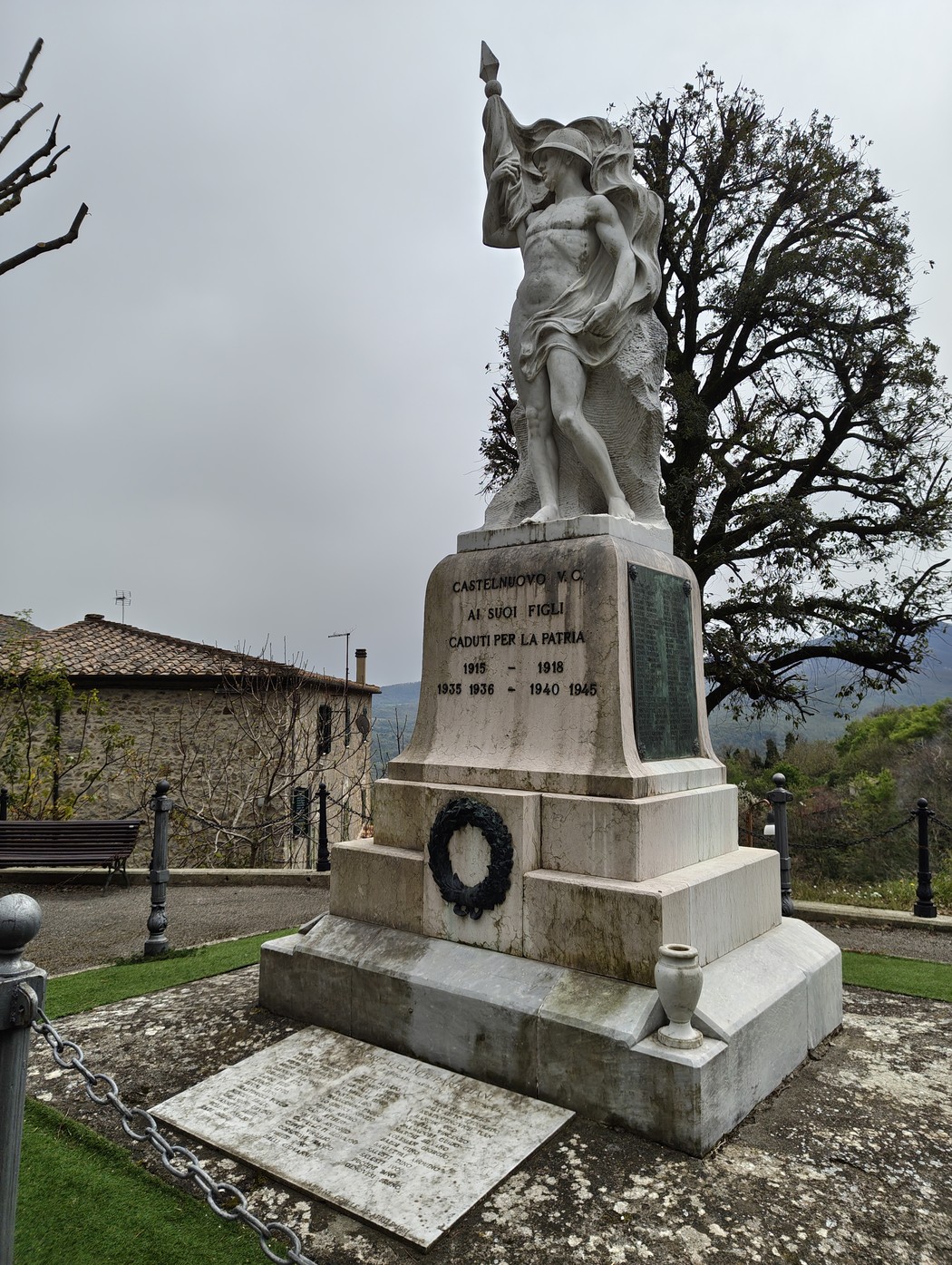 Monumento ai caduti con statua e dedica commemorativa in un paesaggio verde e collinare.