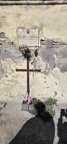 Croce in legno decorata con fiori, posizionata su un muro in rovina con una targa commemorativa.