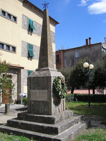 Monumento ai caduti, obelisco commemorativo circondato da vegetazione e adornato da una corona d'alloro.