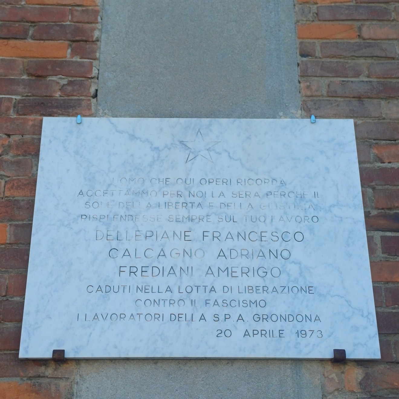 Lapide marmorea con stella e iscrizione, fissata su pilastro tra due pareti in mattoni.